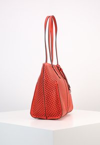 Sac fourre-tout en cuir perforé rouge avec des accents noirs, doté de poignées doubles et d'une fermeture éclair. Surface texturée avec des découpes distinctives.