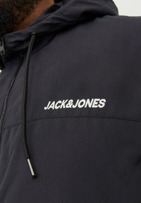 Black hooded jacket with a smooth texture, featuring a white "JACK&JONES" logo on the chest and adjustable drawstrings on the hood.