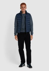 Navy puffer vest over een grijze sweatshirt, zwarte corduroy broek en zwarte sneakers met witte strepen. Casual outfit met een rits aan de voorkant.