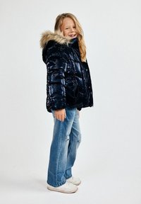 Napušena mornarica puffer jakna s kapuljačom od umjetnog krzna, u kombinaciji s svijetloplavim trganim trapericama i bijelim tenisicama, stoji protiv bijele pozadine.