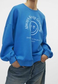 Sudadera azul con cuello redondo, corte oversize y puños acanalados. Presenta texto gráfico blanco y un diseño de cara sonriente en el frente.