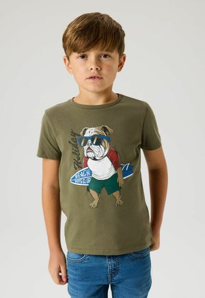 Boy with brown hair wearing olive green t-shirt featuring a bulldog in sunglasses holding a surfboard, paired with blue jeans, standing against plain background.