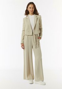 Beige getailleerde cropped blazer met dubbele knopen, gecombineerd met hoogwaardige beige wijde broek. Heeft een zachte, gladde textuur en witte sneakers.