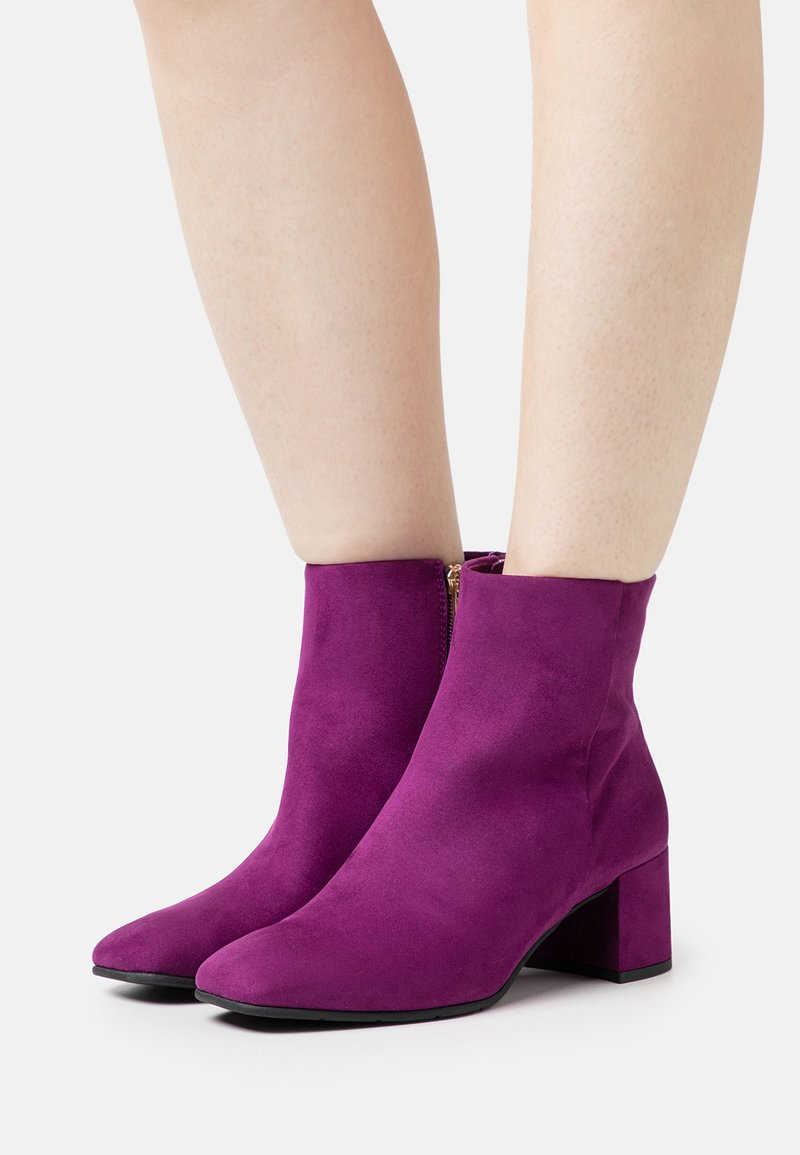 Bottes cheville en daim violet avec talon bloc, bout pointu et détail de fermeture éclair latérale, posées sur un sol gris clair.
