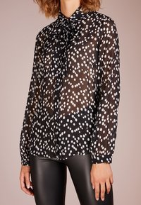 Chemisier noir transparent à pois blancs, à manches longues, avec une fermeture à boutons et un nœud au cou, associé à des leggings noirs brillants.