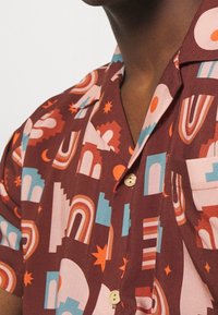 Lyhythihainen paita, jossa on ruskea ja koralli geometrinen kuvio, joka sisältää kaaria, tähtiä ja muotoja. Paita on valmistettu kevyestä kankaasta ja siinä on napitusyksityiskohtia.