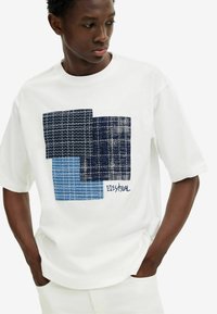 Camiseta de algodón blanca con un diseño de patchwork geométrico en azul y marino. El gráfico incluye acentos cosidos y el nombre de la marca "Desigual".