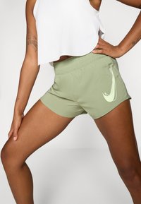 Pantaloni sportivi verdi con una fascia elastica in vita, caratterizzati da un logo swoosh bianco sulla coscia sinistra e un tessuto liscio e leggero.