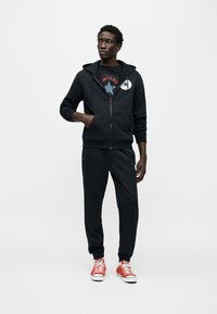 Μαύρη ζακέτα με φερμουάρ και λευκό λογότυπο, συνδυασμένη με μαύρο παντελόνι και κόκκινα αθλητικά παπούτσια. Λείο ύφασμα, casual σχεδιασμός, αθλητική εφαρμογή.