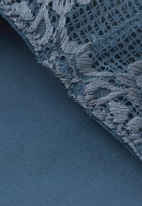Blå tyg med en slät, matt textur nedtill och en utsmyckad, mönstrad spetskant med en glansig yta längs kanten.