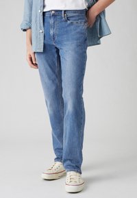 Personne portant un jean bleu clair, des baskets blanches, une chemise blanche et une chemise en denim bleu clair déboutonnée, avec une main dans la poche.
