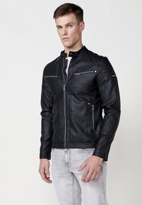 Chaqueta de cuero sintético negro con cuello alto, cierres frontales de cremallera y múltiples bolsillos con cremallera. Presenta acentos acolchados en los hombros.