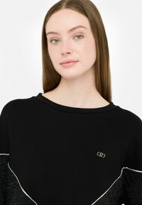 Zwarte sweatshirt met een ronde halslijn, voorzien van een contrasterend sprankelend zwart-wit patroon en een klein gouden logo detail op de borst.