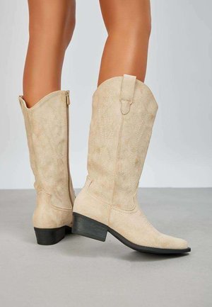Bottes de cowboy en daim beige avec un bout pointu, des motifs embossés subtils et un talon bas empilé. Fermeture éclair latérale pour un enfilage facile.