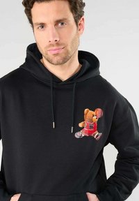 Zwarte hoodie met verstelbare touwtjes, met een teddybeerprint in een rood sportshirt die een basketbal vasthoudt. Zachte stof met een relaxte pasvorm.