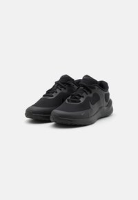 Chaussures de sport noires avec une tige en mesh, bout arrondi et semelle texturée. Les caractéristiques incluent un système de laçage et un logo discret sur le côté.