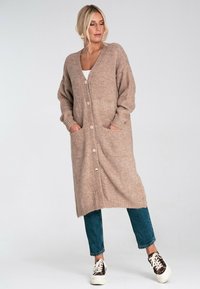 Kobieta ubrana w długi beżowy kardigan zapinany na guziki z kieszeniami, niebieskie dżinsy i trampki w panterkę, stojąca na tle jednolitego białego tła.