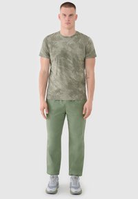 Un homme est debout, face à l'avant, portant une chemise à manches courtes camouflage verte, un pantalon vert olive et des baskets grises et blanches sur un fond uni.