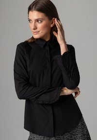 Black long-sleeve shirt with a collared neckline and ruffled cuffs, made of smooth fabric. The fit is tailored, featuring a curved hem.