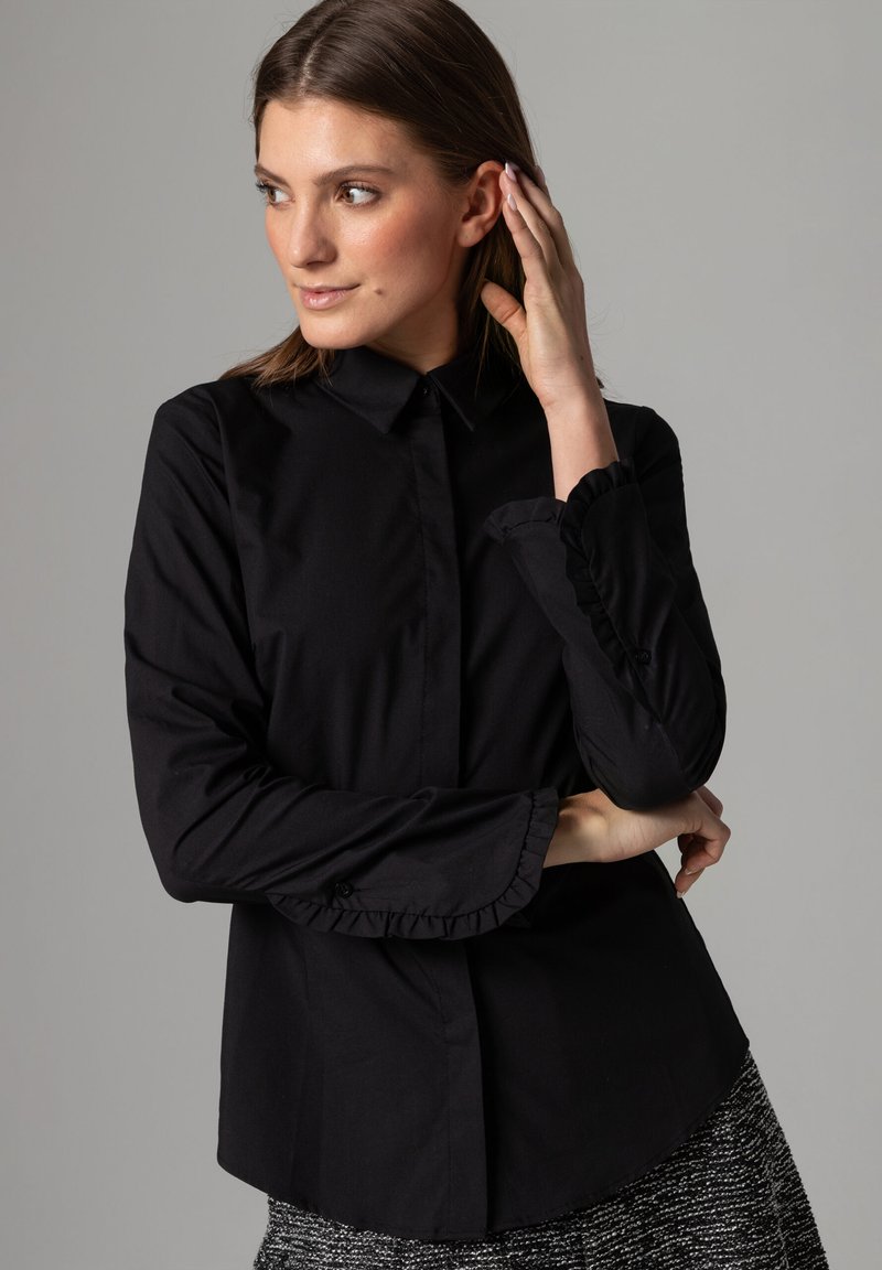 Black long-sleeve shirt with a collared neckline and ruffled cuffs, made of smooth fabric. The fit is tailored, featuring a curved hem.