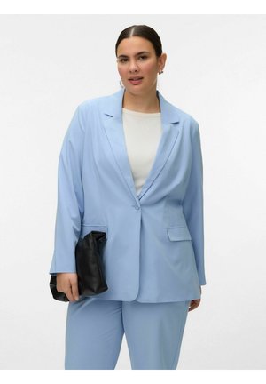 Nő világoskék öltönyben és fehér felsőben, fekete kézitáskát tartva, egyszínű fehér háttér előtt.