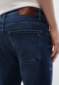 Tamno plave traperice s kožnom BOSS oznakom na stražnjoj strani struka, s standardnim džepovima i suptilnim detaljima šavova.