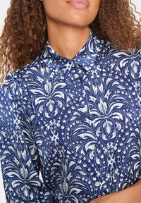 Camicia blu navy con motivo floreale bianco, dotata di colletto e chiusura con bottoni. Il materiale sembra liscio con una leggera lucentezza.