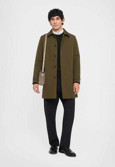 Abrigo verde oliva con botones en la parte delantera, llevado sobre un suéter oscuro y una camisa, combinado con pantalones oscuros y zapatos negros, llevando un bolso pequeño.