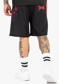 Sorte shorts med røde og lilla grafiske accenter, elastisk talje, og en baglomme. Sammen med hvide sneakers og stribede sokker.