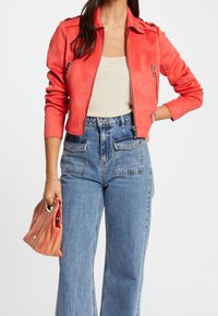 Czerwona kurtka z sztucznego zamszu z zamkami, krótki fason, z naramiennikami, w połączeniu z niebieskimi dżinsami o wysokim stanie i wzorzystą torebką.