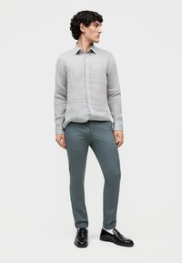 Młody mężczyzna stojący, ubrany w jasnoszarą koszulę na guziki, turkusowe wąskie spodnie z mankietami oraz czarne skórzane loafersy, patrzący w lewo.