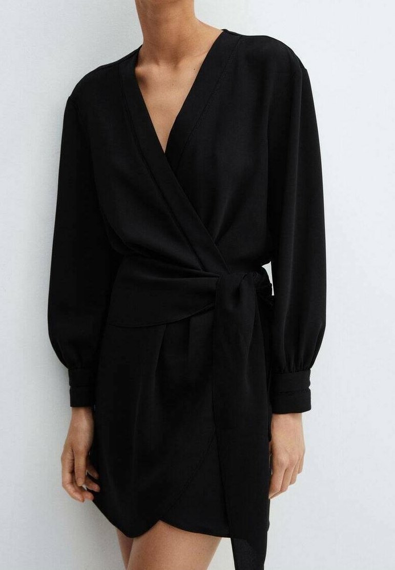 Femme portant une robe portefeuille noire à manches longues avec un décolleté en V profond et une ceinture nouée à la taille, sur un fond clair uni.