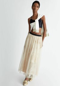 Top corto nero con un maglione bianco lavorato a maglia drappeggiato sulle spalle, abbinato a una gonna leggera e trasparente di tonalità dorata. Calzature con accenti dorati.