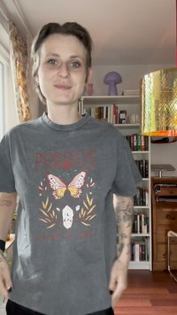 T-shirt de algodão cinza com um corte solto, apresentando gráficos coloridos de borboletas e cristais com o texto "POSITIVO" e "SEMPRE NA ALMA."
