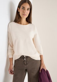 Helle beige Strickjacke mit weitem Ausschnitt und Dreiviertelärmeln, kombiniert mit dunkelbraunen Hosen, die mit zwei Knöpfen geschlossen werden.