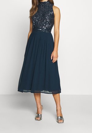 Sleeveless navy midi dress with sequined high-neck bodice and flowy chiffon skirt, paired with silver open-toe heels.