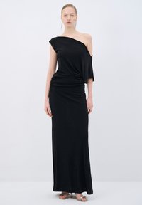 Sort, asymmetrisk, off-shoulder maxikjole med rynkede detaljer og blødt stof. Modellen bærer metalliske, strappy højhælede sko.
