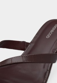 Κοντινό πλάνο ενός σκούρου καφέ δερμάτινου σανδαλιού με δύο ραμμένες λουρίδες που διασταυρώνονται πάνω στο πέλμα, σε λευκό φόντο.