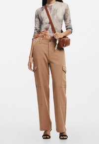 Pantaloni cargo beige con tasche laterali, abbinati a una maglietta a maniche lunghe con motivo grigio e arancione. Gli accessori includono una borsa a tracolla marrone.