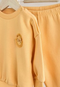 Gul sweatshirt med ribbad halsringning och axelknappar, med ett lejonmotiv, parat med matchande sweatpants med elastisk midja.