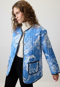 Plisirana jakna u plavoj boji s bijelim cvjetnim uzorkom, koja ima okrugli ovratnik, dva prednja džepa i crne obrube duž rubova.