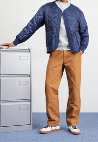 Homme portant une veste matelassée bleue, un sweat-shirt blanc, un pantalon marron et des baskets blanches, debout avec une main posée sur un classeur gris.