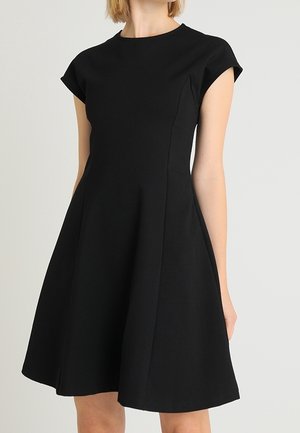 Frau trägt ein kurzärmeliges, knielanges schwarzes A-Linien-Kleid mit figurbetontem Oberteil und rundem Ausschnitt vor einem schlichten Hintergrund.