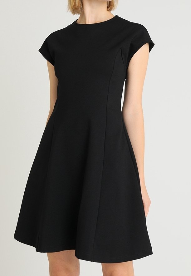 Femme portant une robe noire trapèze à manches courtes, longueur genou, avec un corsage ajusté et un col rond sur un fond uni.