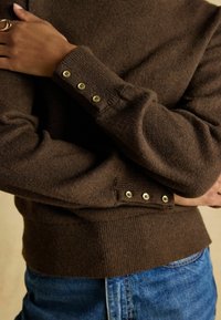 Brauner Pullover mit gerippter Textur, ausgestattet mit goldenen Knöpfen an den Ärmeln. Kombiniert mit blauer Denim, die ein figurbetontes Design zeigt.