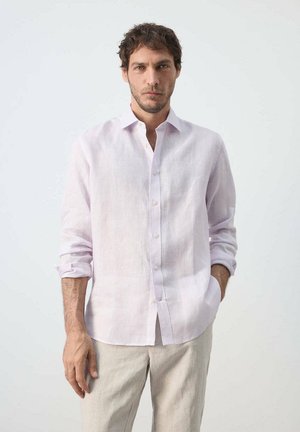 Homme aux cheveux courts et bouclés portant une chemise rose clair à boutons et un pantalon beige, debout avec une main dans la poche devant un fond uni.