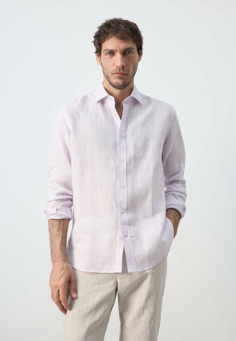 Homme aux cheveux courts et bouclés portant une chemise rose clair à boutons et un pantalon beige, debout avec une main dans la poche devant un fond uni.