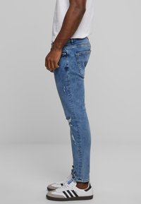 Leblített kék skinny farmernadrág, kopott részletekkel, zsebekkel és övhurkokkal. Fehér tornacipővel és fekete csíkokkal párosítva.