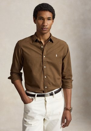 Jeune homme aux cheveux bouclés portant une chemise marron à manches longues boutonnée et un pantalon blanc avec une ceinture noire sur un fond uni.