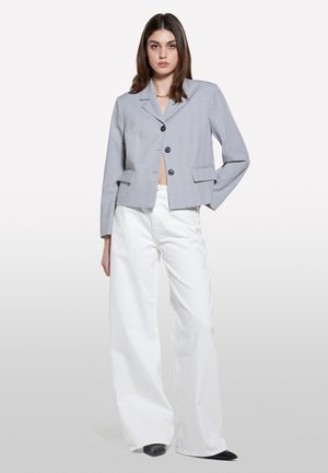 Blazer gris cropped avec un col à revers, deux poches à l'avant et des boutons noirs, associé à un pantalon large blanc et des chaussures à talons noirs.